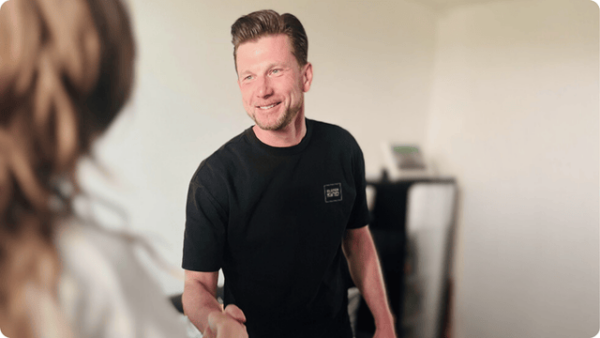 Ein junger Mann begrüßt eine Person mit einem Handschlag und lächelt freundlich dabei.