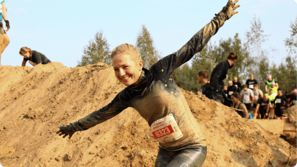 Teilnehmerin eines Hindernislaufs lacht und posiert verschmutzt vor einem Sandhügel während eines Outdoor-Mud-Runs.